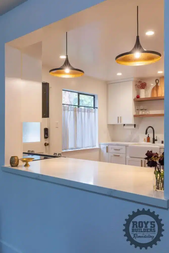 View through a kitchen pass-through of a modern kitchen with white cabinets, a farmhouse sink, black refrigerator, and two black pendant lights.