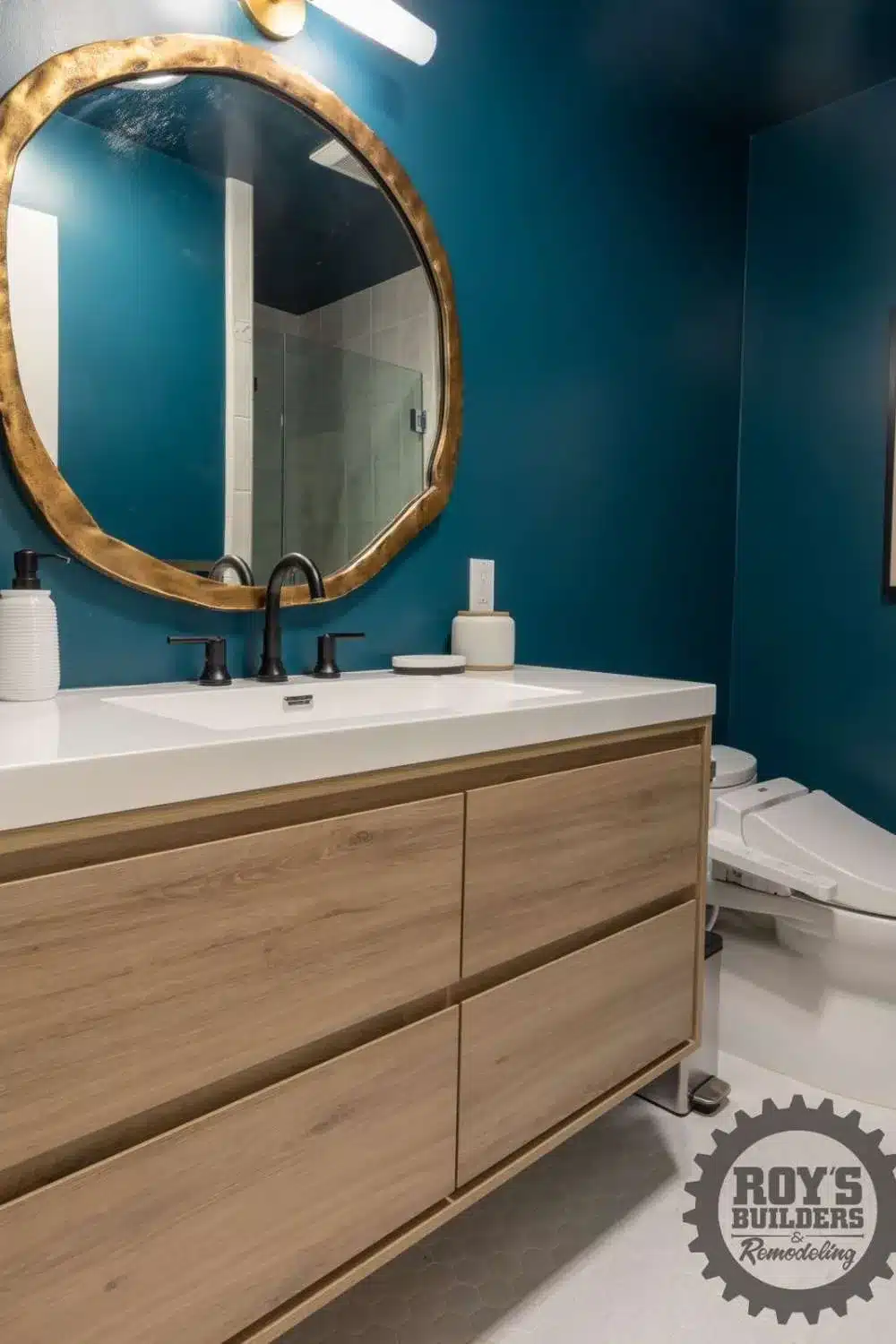 Natural wood vanity with a white countertop and a large gold mirror in a bathroom with dark teal walls