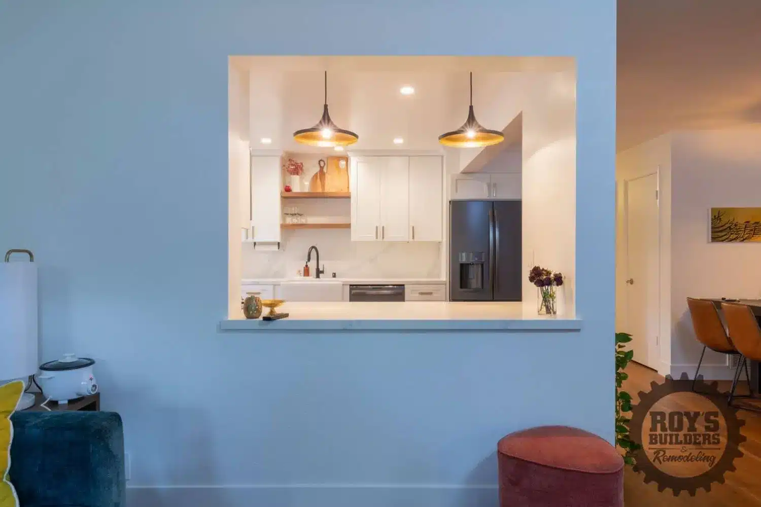View through a kitchen pass-through of a modern kitchen with white cabinets, a farmhouse sink, black refrigerator, and two black pendant lights.