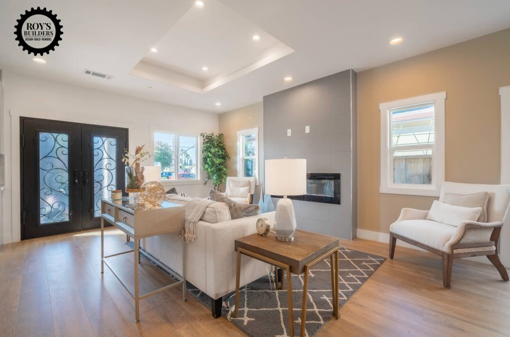 Modern living room remodel by Roy’s Builders featuring wood flooring, a sleek gray fireplace wall, stylish beige furniture, and elegant black double doors with decorative glass.