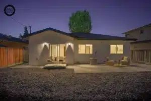 Rear view of a white one-story modern house at night with glowing indoor and outdoor lights, featuring a concrete patio, wicker furniture, and a dark gravel yard.