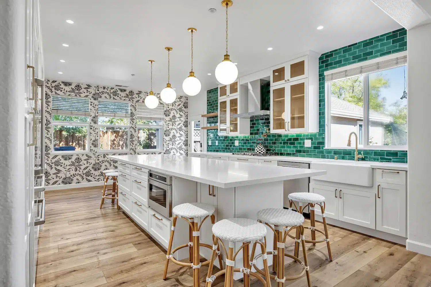 An airy kitchen with white cabinetry, a large white island with gold-trimmed stools, and a vibrant emerald green tiled backsplash.