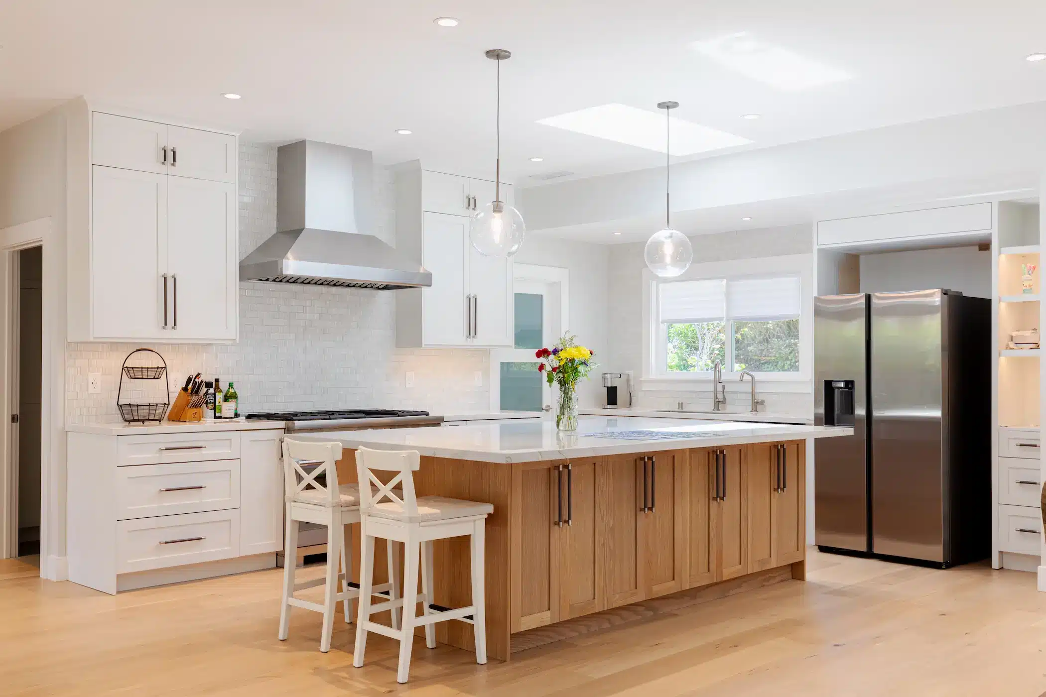An airy kitchen featuring white upper cabinets, a light natural wood island with a white countertop, and elegant glass pendant lights.