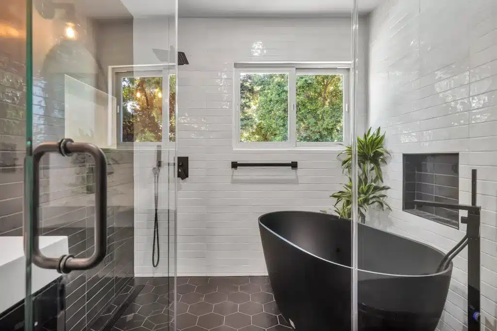 A contemporary bathroom featuring a freestanding matte black oval bathtub, a glass-enclosed shower with white subway tile, and dark hexagonal floor tiles.