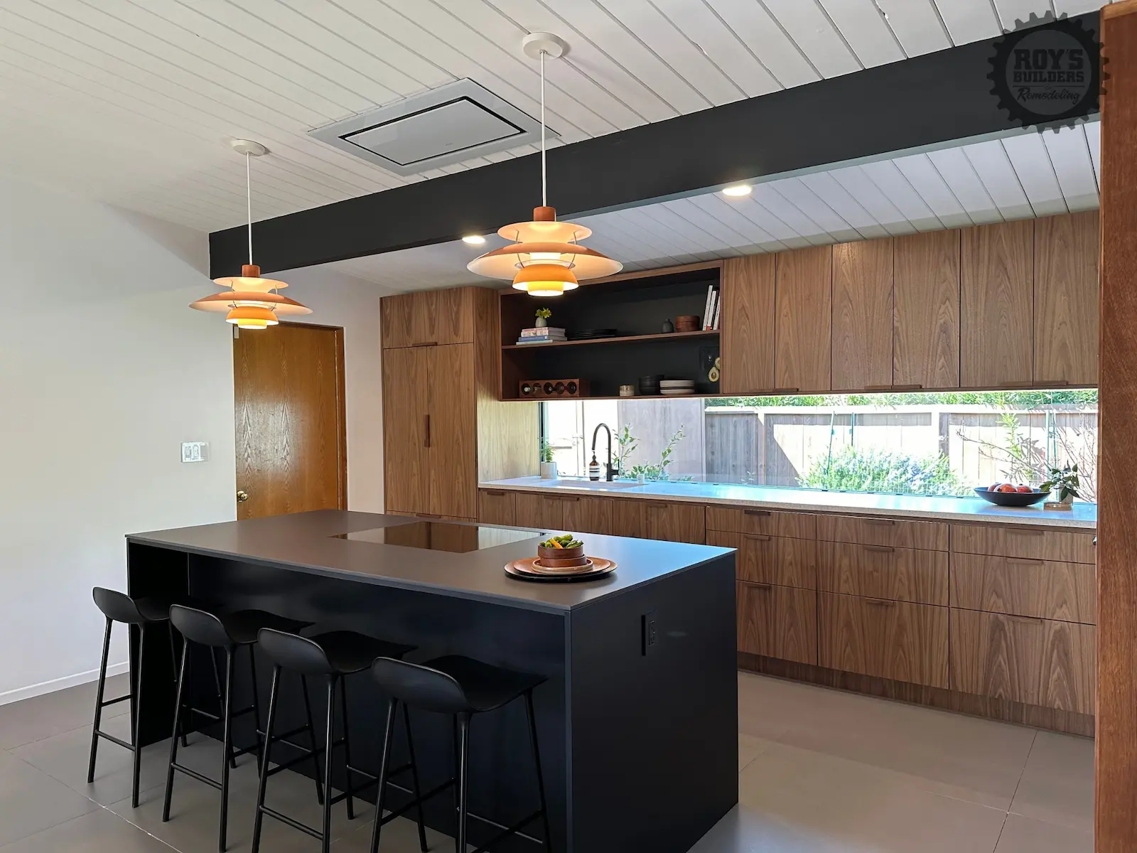 A kitchen with horizontal grain wood cabinets, a black kitchen island, and iconic orange saucer pendant lights hanging from a white slatted ceiling.