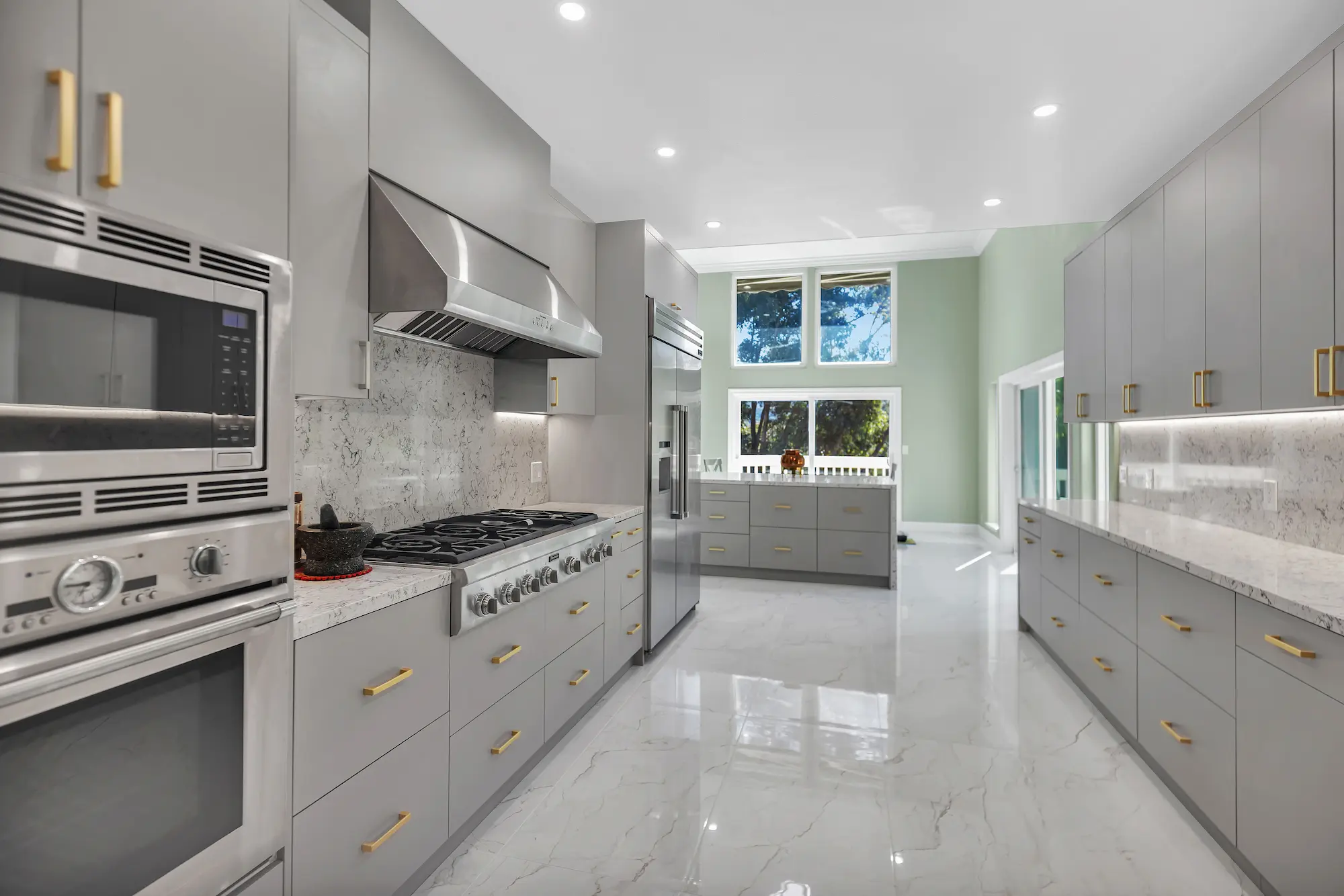 A kitchen featuring sleek light grey cabinetry with gold handles, a large stainless steel gas range, and polished marble flooring.
