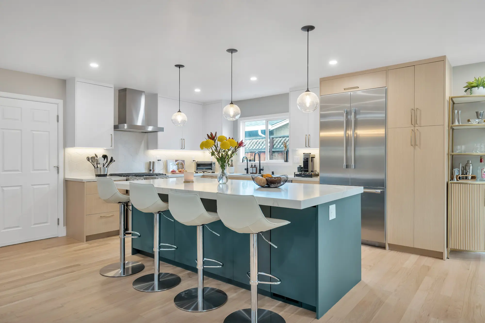 A bright kitchen with white perimeter cabinets, light wood floors, and a large deep teal kitchen island with a white waterfall countertop and four white barstools.