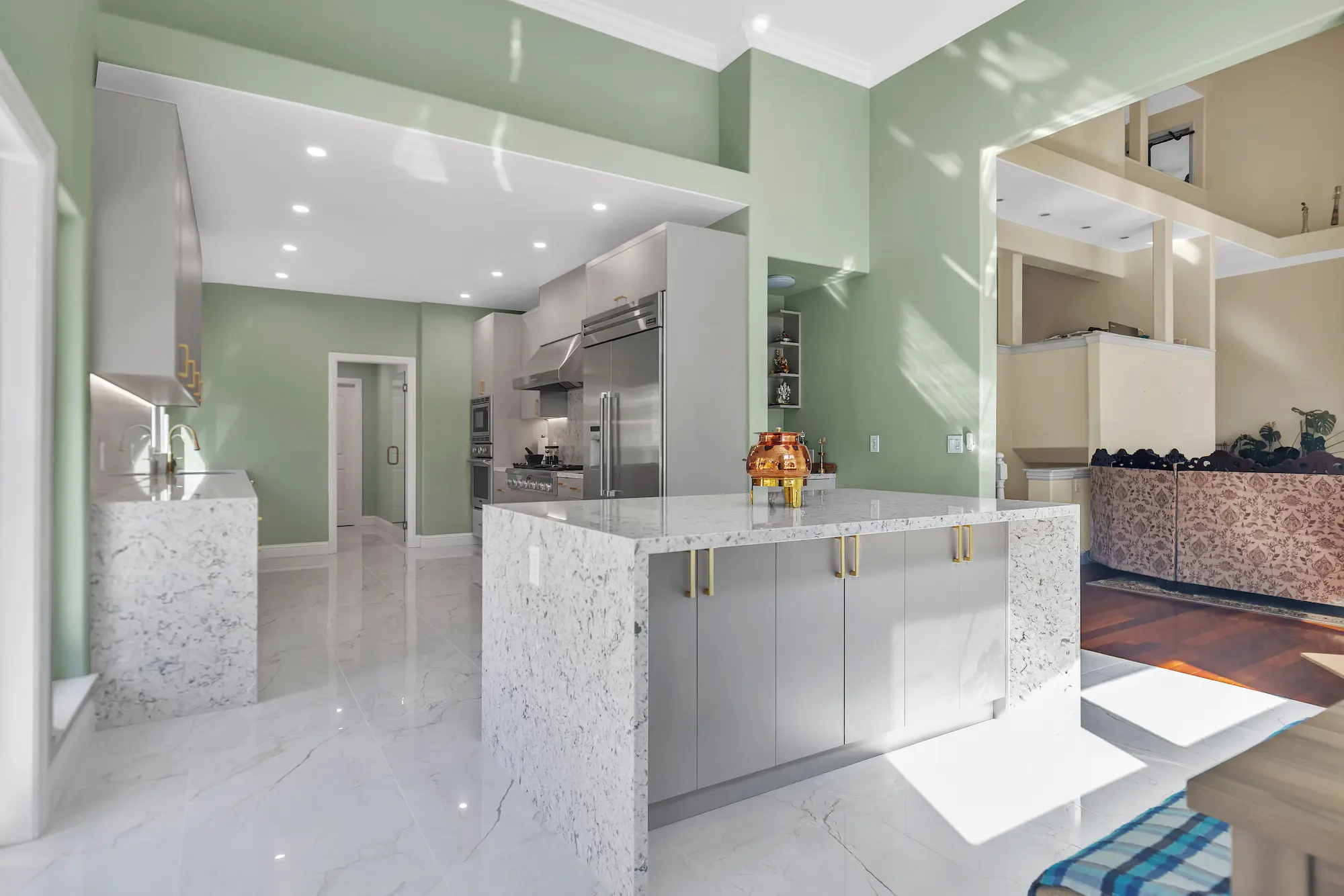 A spacious kitchen with sage green walls, grey cabinetry, and a massive marble-clad waterfall island under bright recessed lighting.