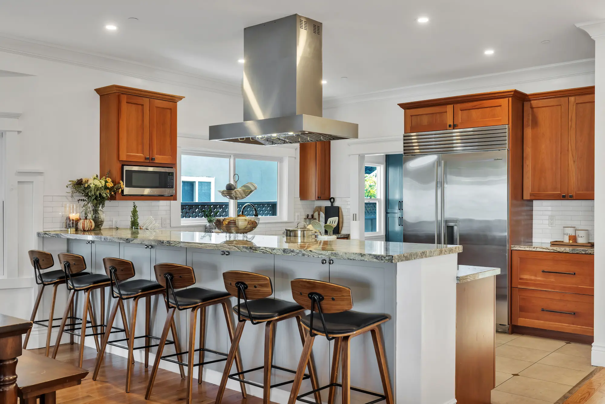 A kitchen featuring warm medium-tone wood cabinets, a large stainless steel refrigerator and range hood, and a white breakfast bar with five wooden barstools.
