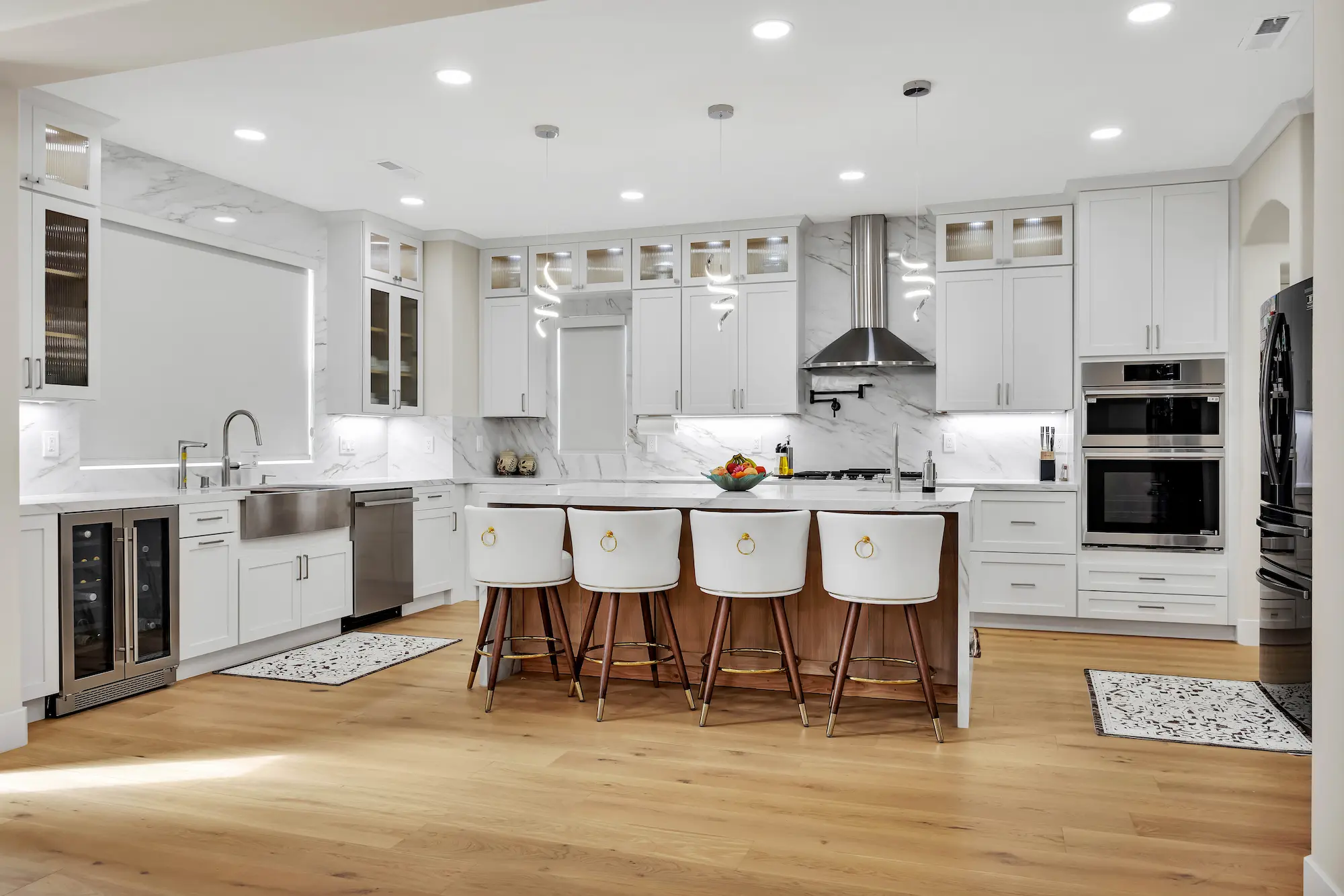Wide shot of a bright kitchen with white cabinetry, light wood floors, integrated stainless steel appliances, and a marble backsplash extending to the ceiling.