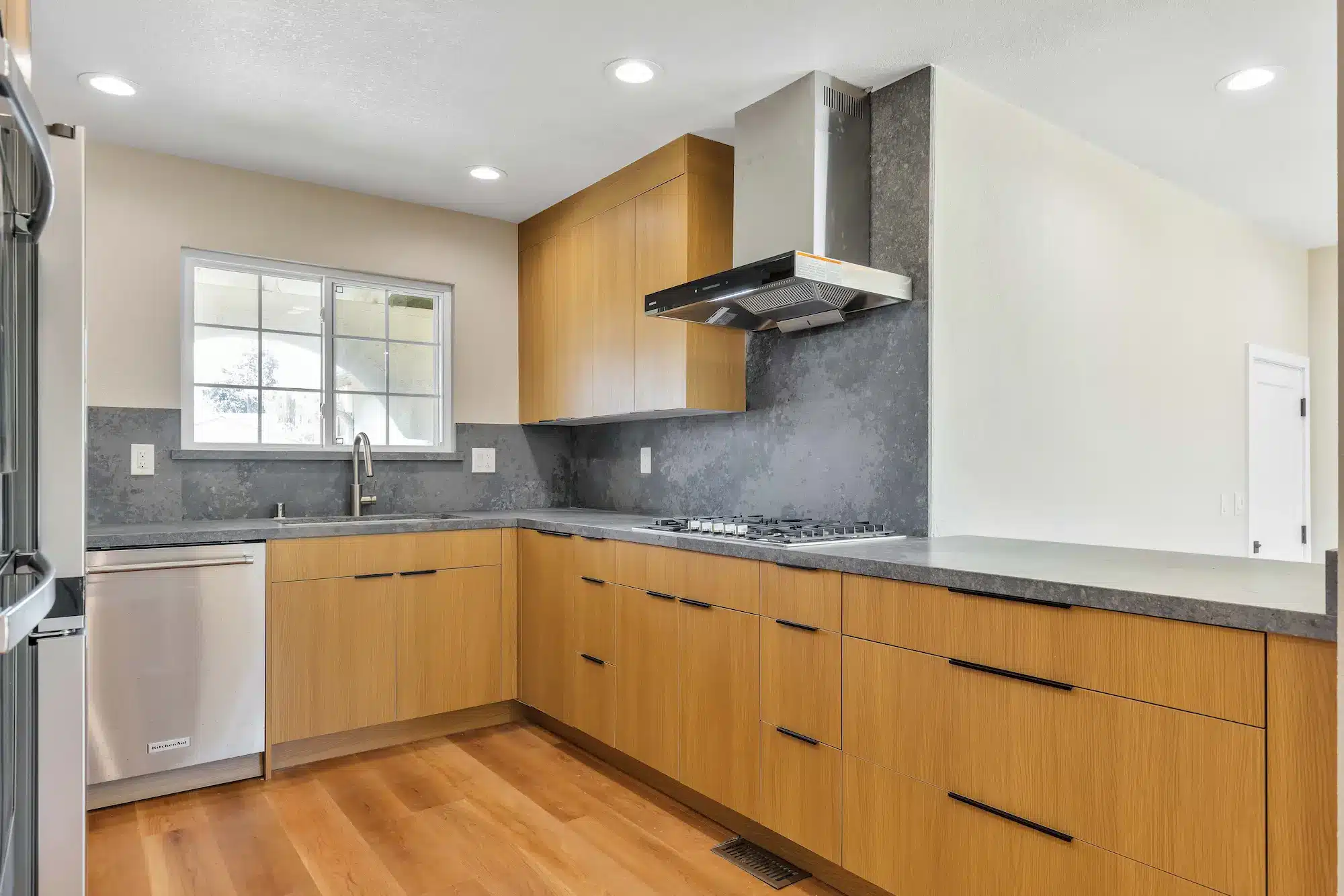 A corner view of a kitchen with wood cabinets, stainless steel dishwasher, and a grey backsplash.