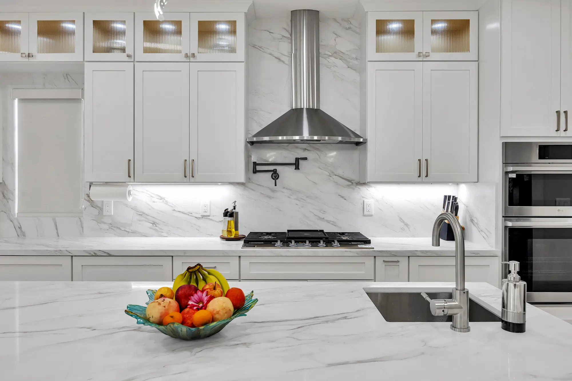 Close-up of a kitchen cooktop and hood with white cabinetry, marble countertops featuring a fruit bowl, and a pot filler faucet installed on the marble backsplash.