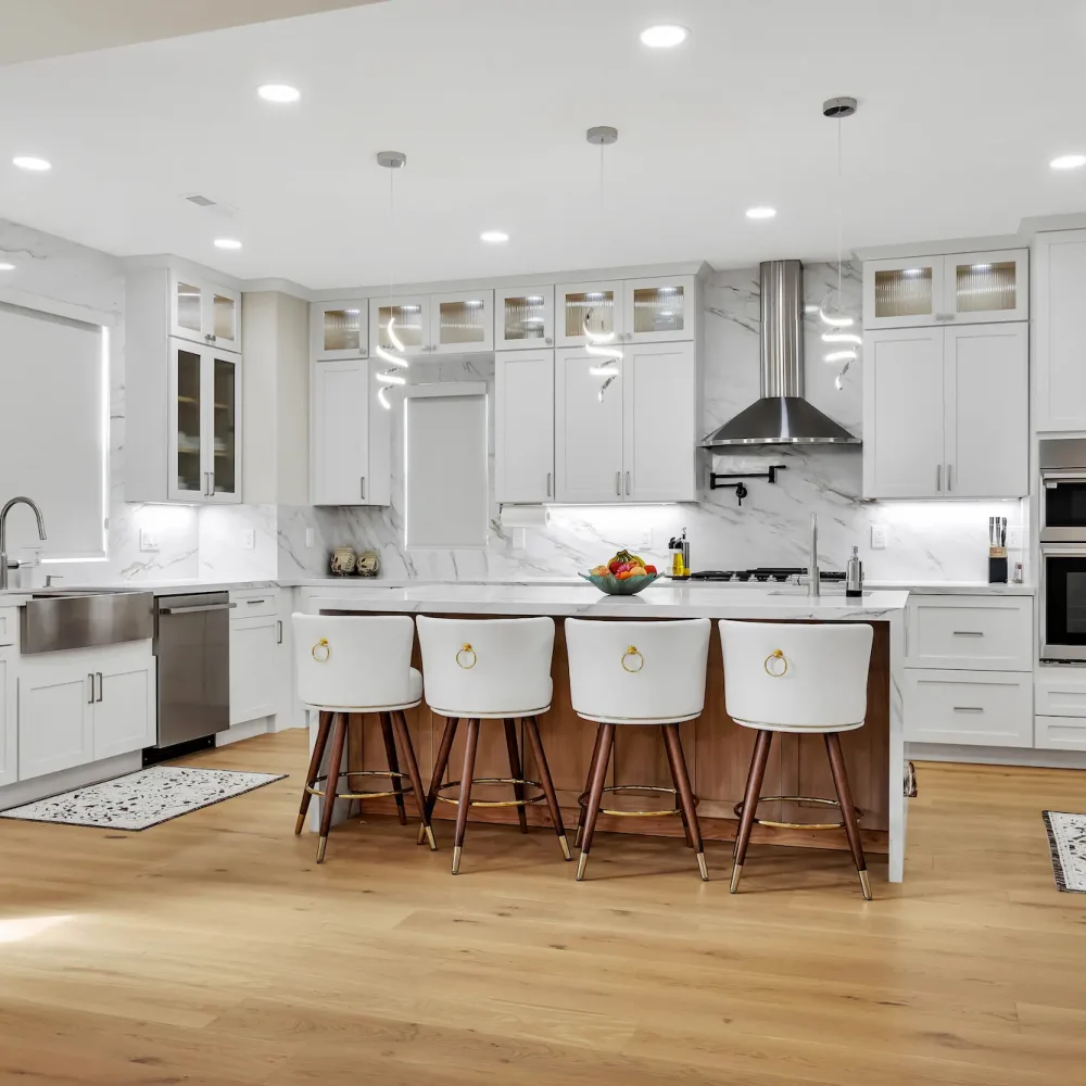Wide shot of a bright kitchen with white cabinetry, light wood floors, integrated stainless steel appliances, and a marble backsplash extending to the ceiling.