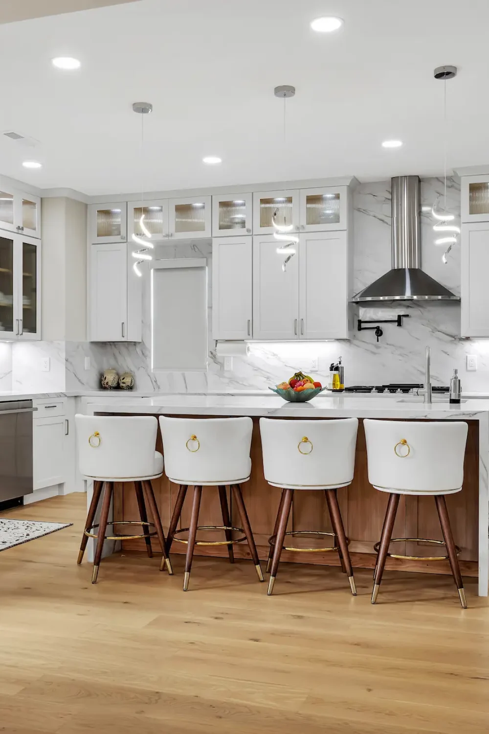 Wide shot of a bright kitchen with white cabinetry, light wood floors, integrated stainless steel appliances, and a marble backsplash extending to the ceiling.