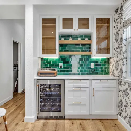 A small auxiliary kitchen station with a wine fridge, green tiles, and floral wallpaper.