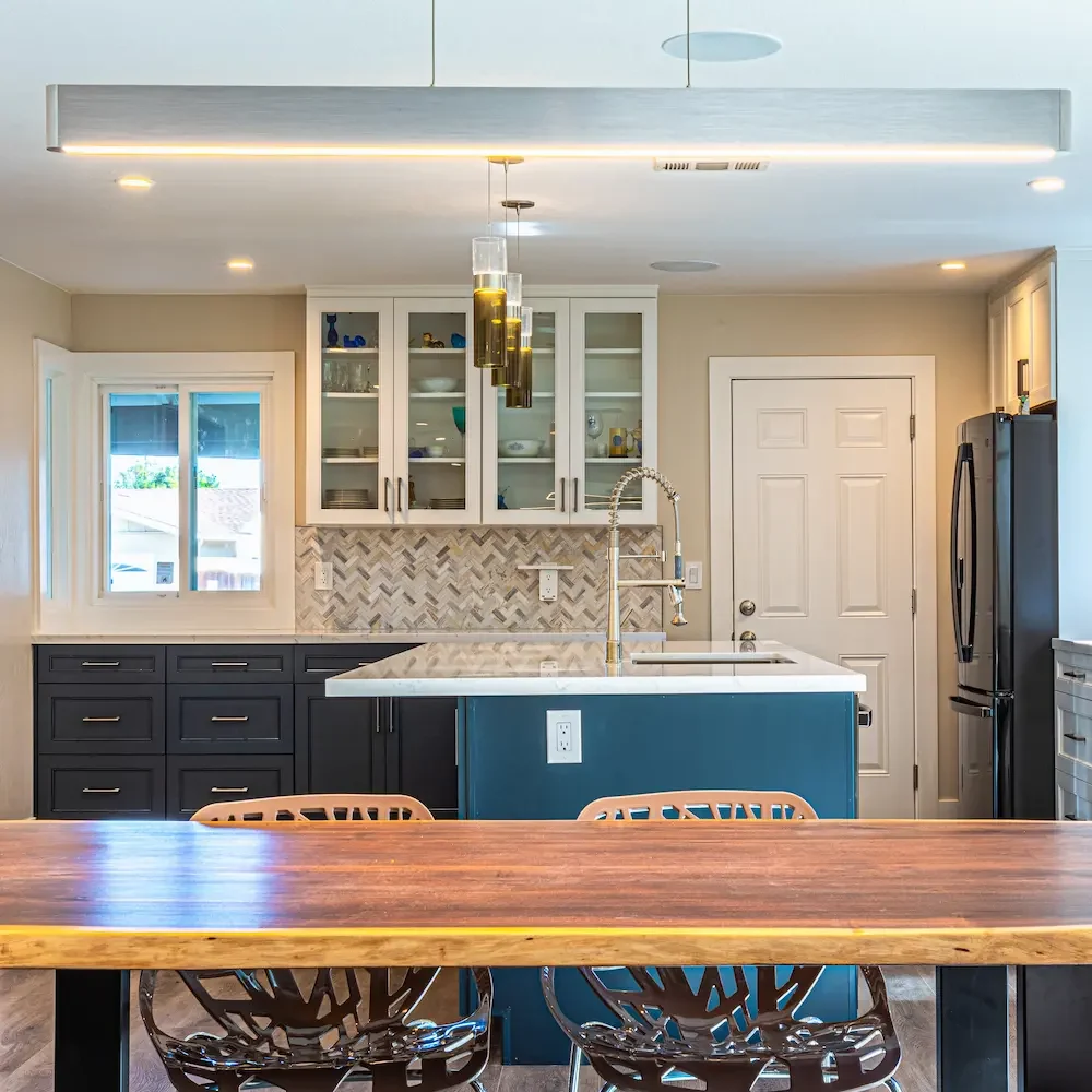 A bright kitchen view from behind a wooden dining table with wire-frame chairs, showing a teal island with a white countertop and a background of dark lower cabinets, a patterned backsplash, and a modern silver linear light fixture hanging from the ceiling.