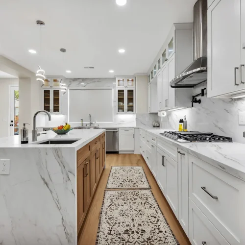 Wide-angle view of a long kitchen with marble countertops, white cabinets, and a runner rug.