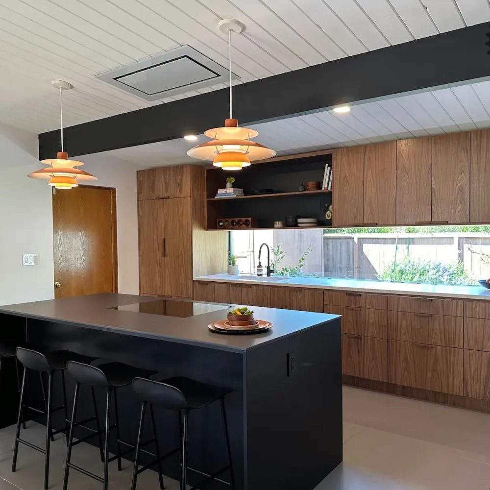 A kitchen with horizontal grain wood cabinets, a black kitchen island, and iconic orange saucer pendant lights hanging from a white slatted ceiling.