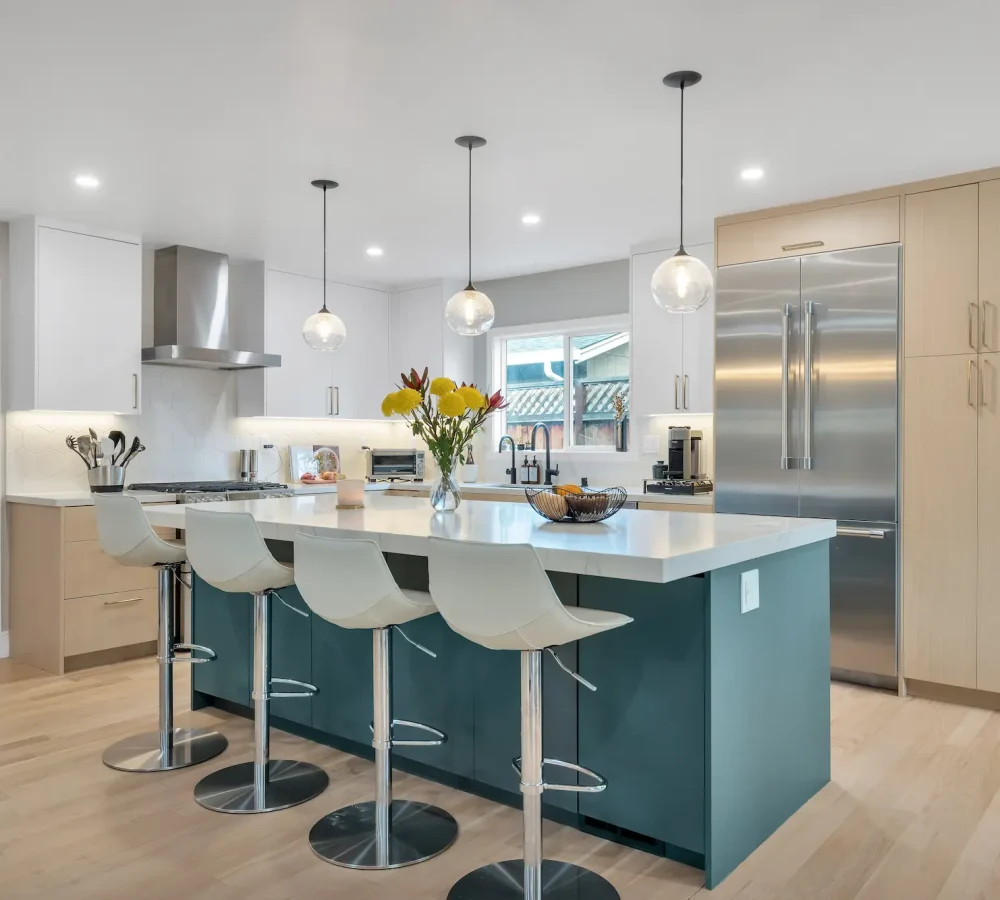 A bright kitchen with white perimeter cabinets, light wood floors, and a large deep teal kitchen island with a white waterfall countertop and four white barstools.