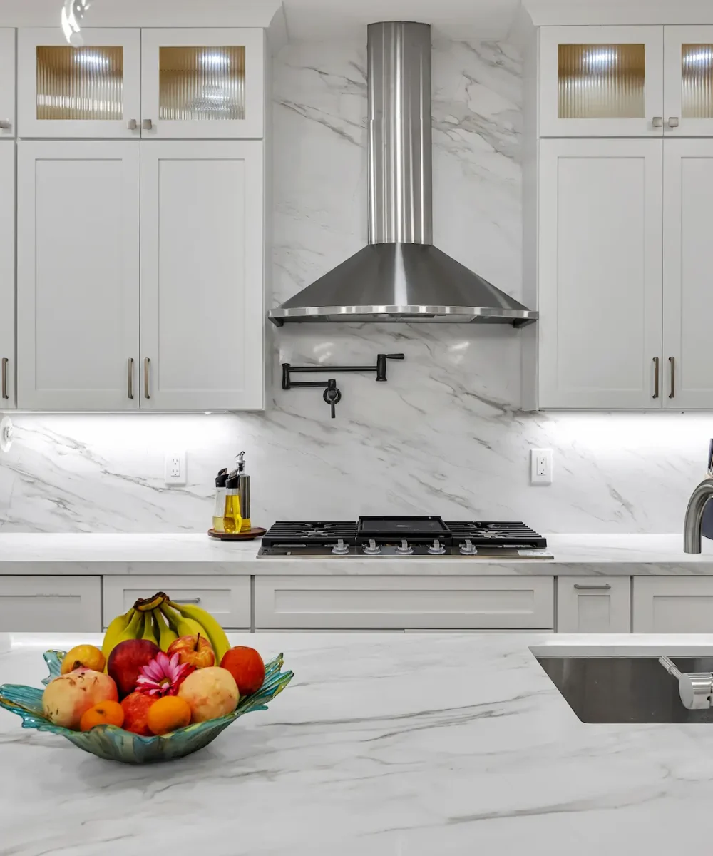 Close-up of a kitchen cooktop and hood with white cabinetry, marble countertops featuring a fruit bowl, and a pot filler faucet installed on the marble backsplash.