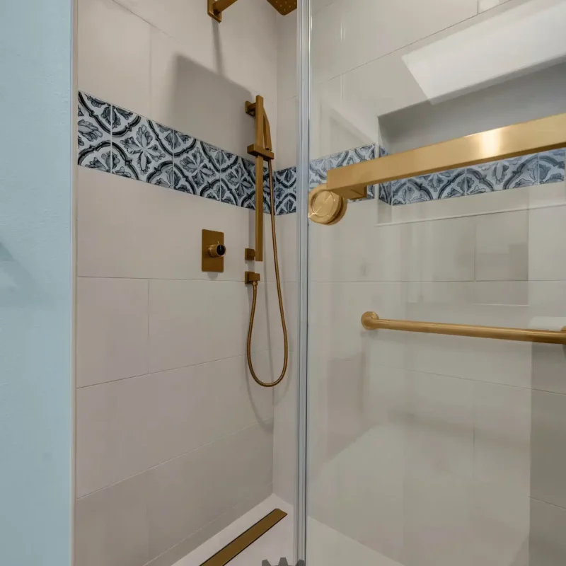Walk-in shower featuring white tiles, a decorative blue pattern accent band, and gold fixtures.