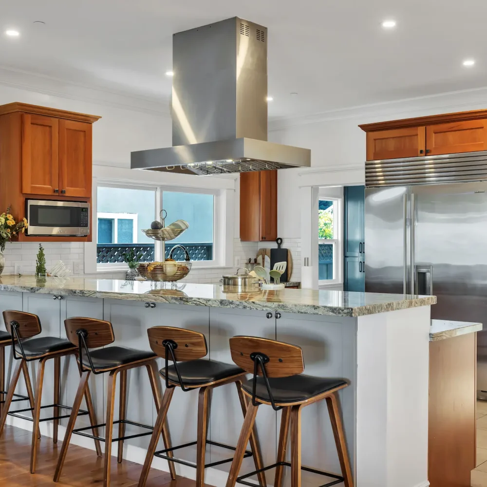 A kitchen featuring warm medium-tone wood cabinets, a large stainless steel refrigerator and range hood, and a white breakfast bar with five wooden barstools.
