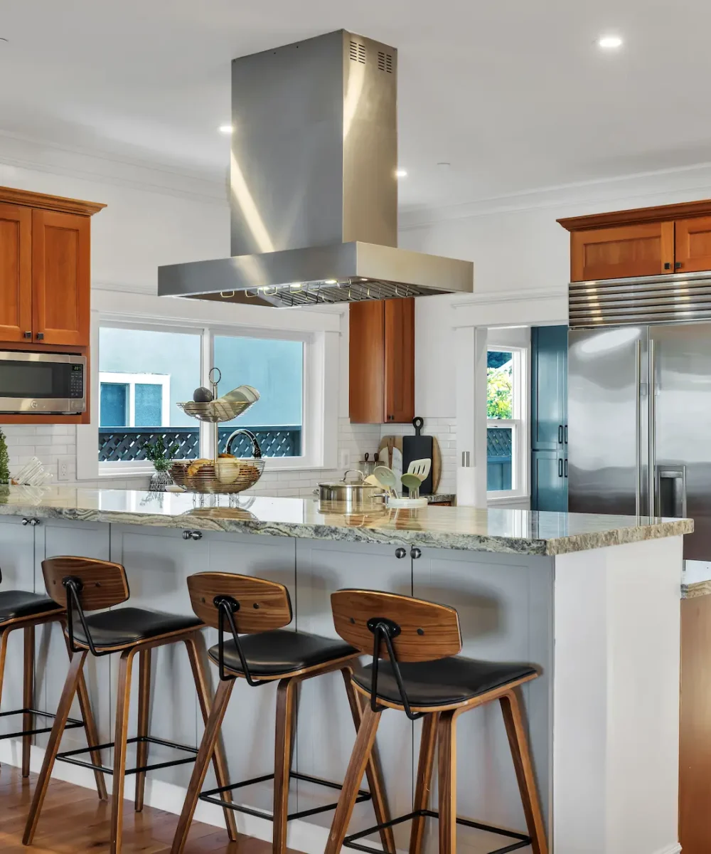 A kitchen featuring warm medium-tone wood cabinets, a large stainless steel refrigerator and range hood, and a white breakfast bar with five wooden barstools.