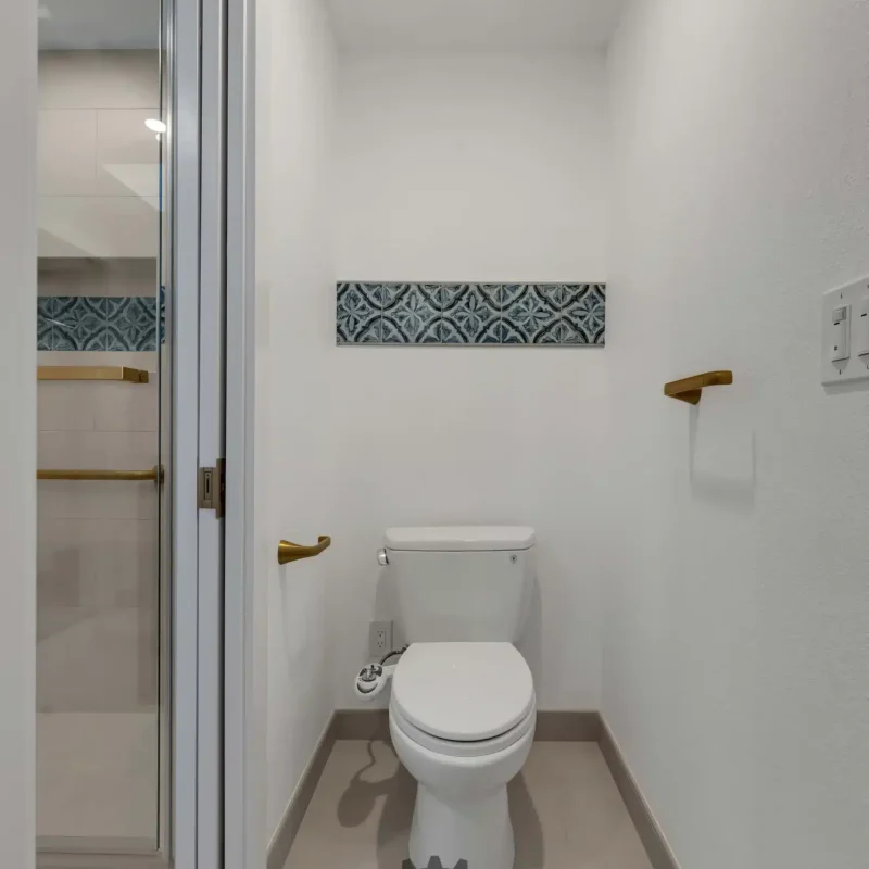 A small restroom showing a toilet, white walls, and a glimpse into a tiled shower.